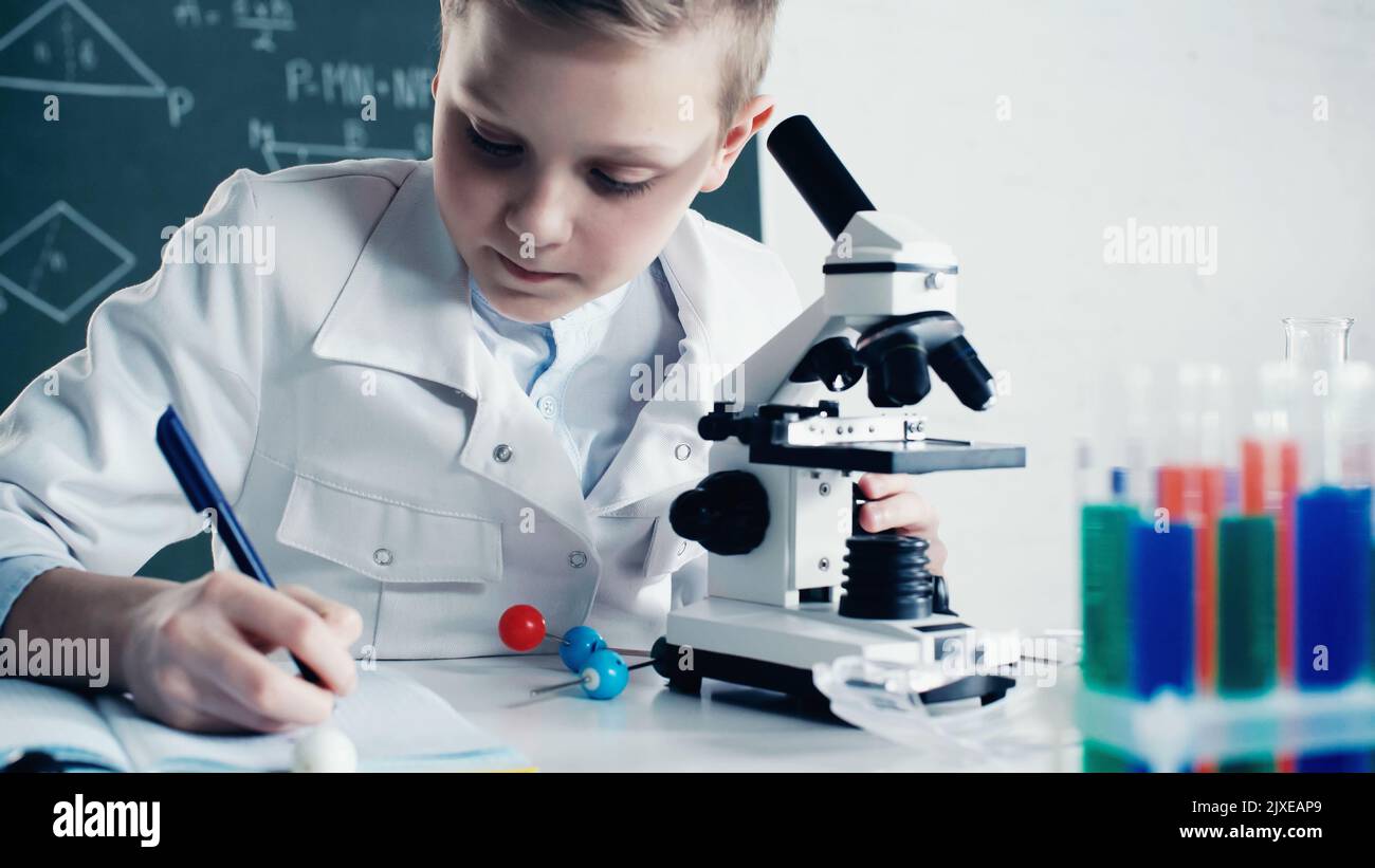 scolaro in cappotto bianco facendo appunti vicino al microscopio durante la lezione in classe, immagine di scorta Foto Stock