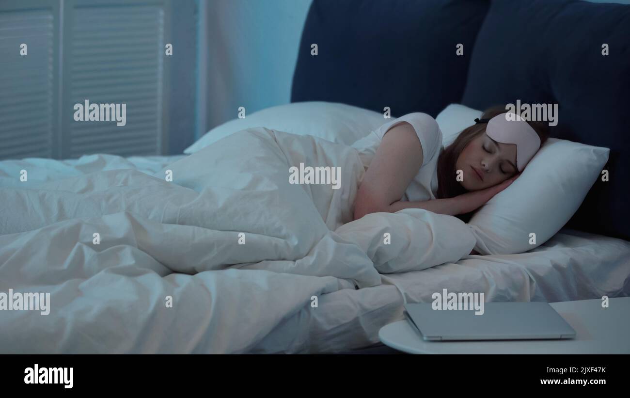 Donna in maschera che dorme sul letto vicino al portatile sul comodino, immagine stock Foto Stock