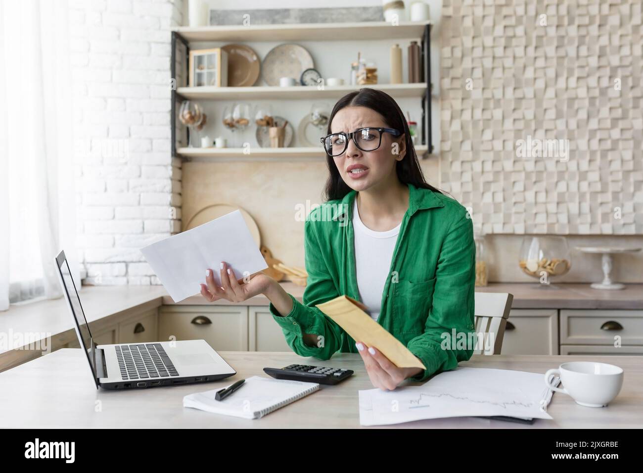 Mutuo ipotecario. Giovane donna arrabbiata e sconvolta che tiene in mano un conto di affitto. Ha ricevuto una lettera sul debito. Sittin g in cucina a casa al tavolo con un computer portatile in bicchieri e una giacca verde Foto Stock