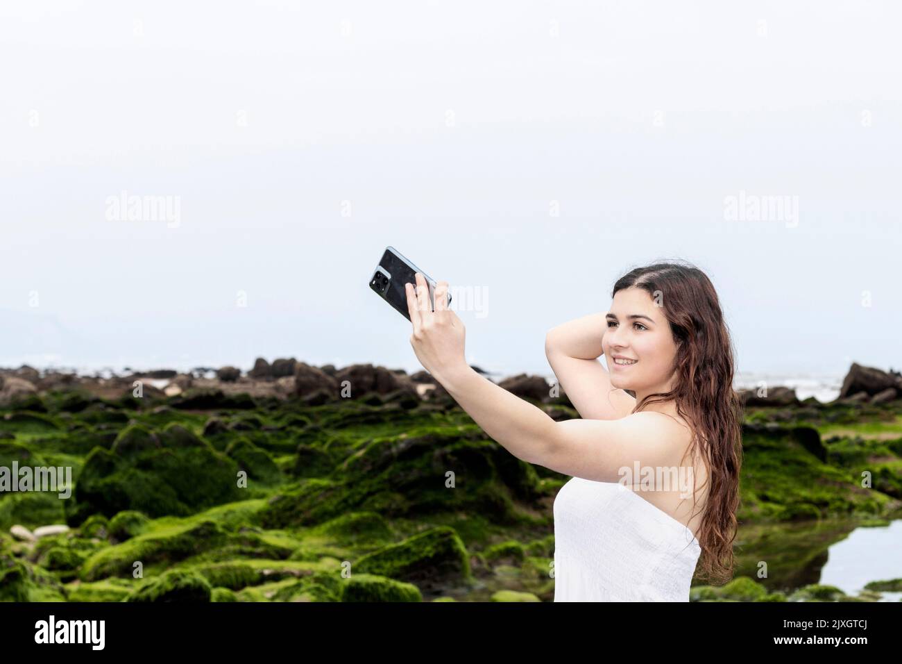 giovane donna brunetta in abito bianco che prende un selfie sulla costa Foto Stock