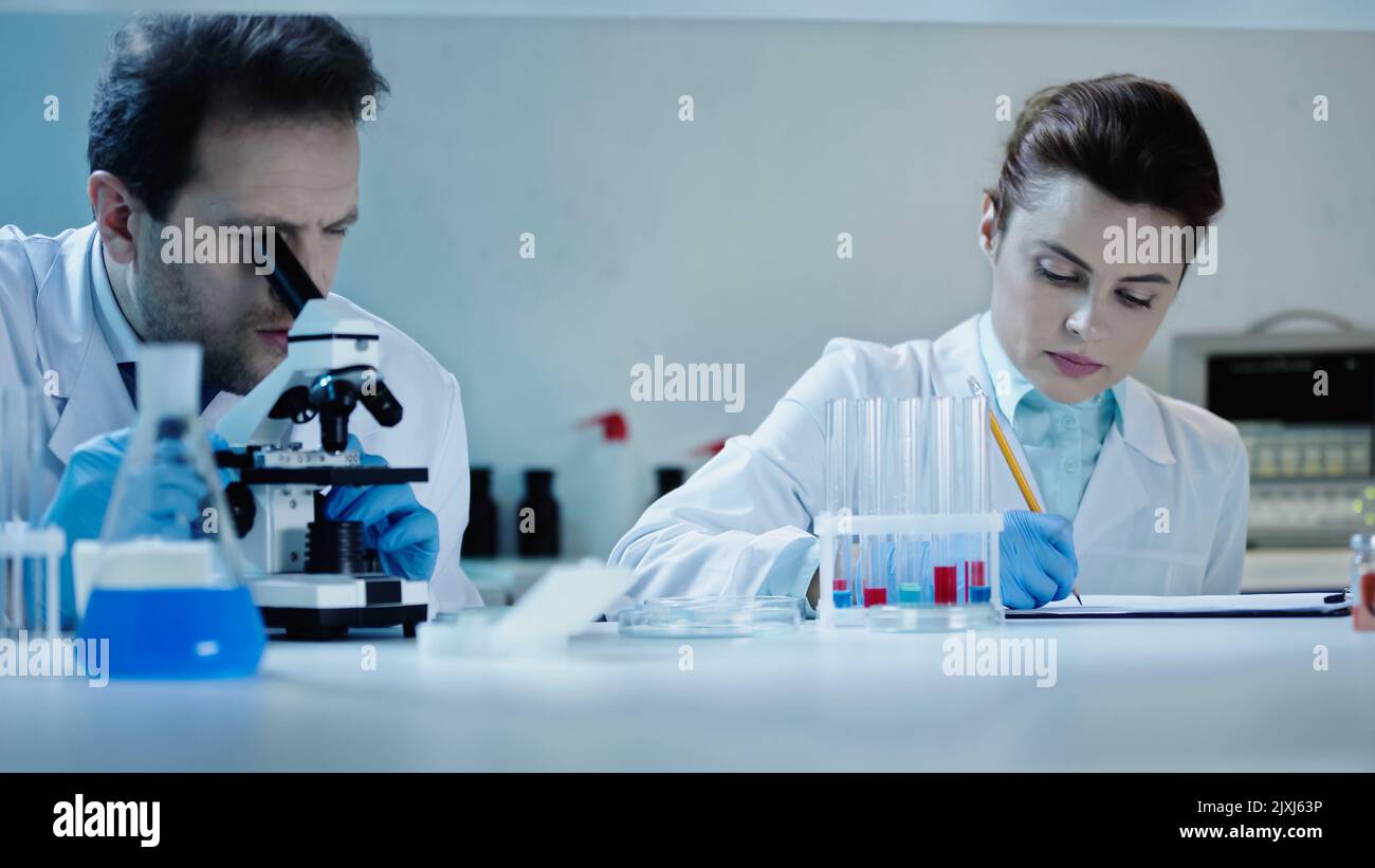 scienziato in cappotto bianco guardando attraverso il microscopio vicino alla scrittura del collega risultato in laboratorio, immagine di scorta Foto Stock