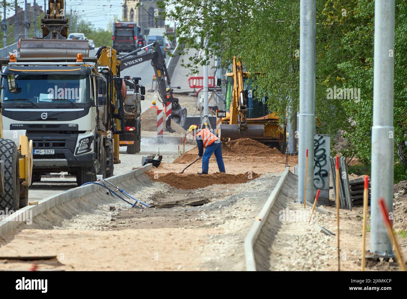 RIGA, LETTONIA - 13 MAGGIO 2022: Riparazione stradale in via Deglava. Un lavoratore in uniforme scava sabbia con una pala. Lavoro duro. Macchine da costruzione pesanti in piedi accanto. Foto Stock
