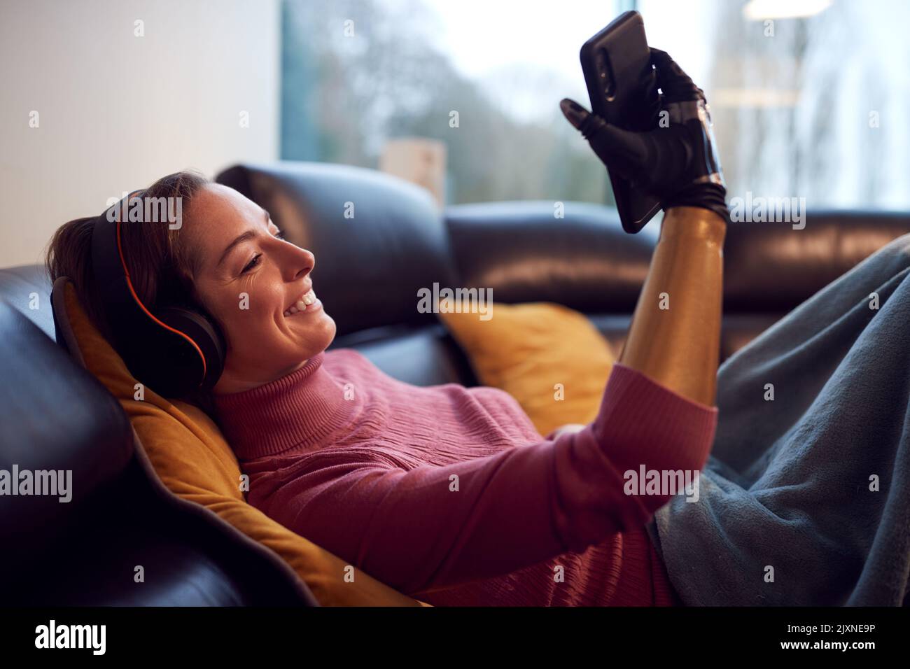 Donna con braccio protesico indossando cuffie senza fili ascolto di musica sul telefono cellulare sul divano Foto Stock