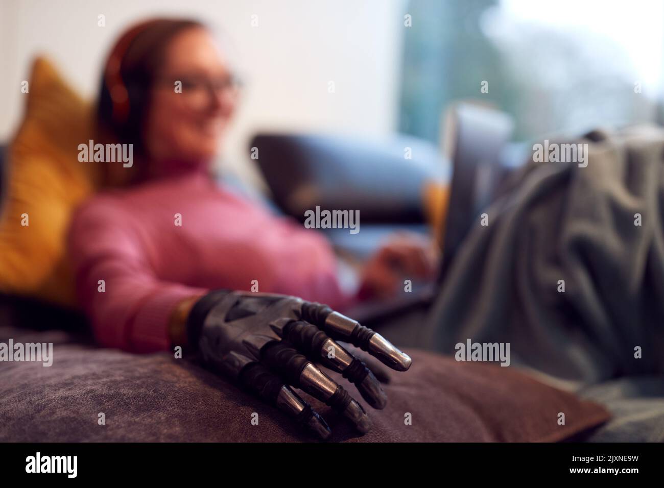 Donna con braccio protesico che indossa cuffie wireless e che lavora su un computer portatile sul divano a casa Foto Stock