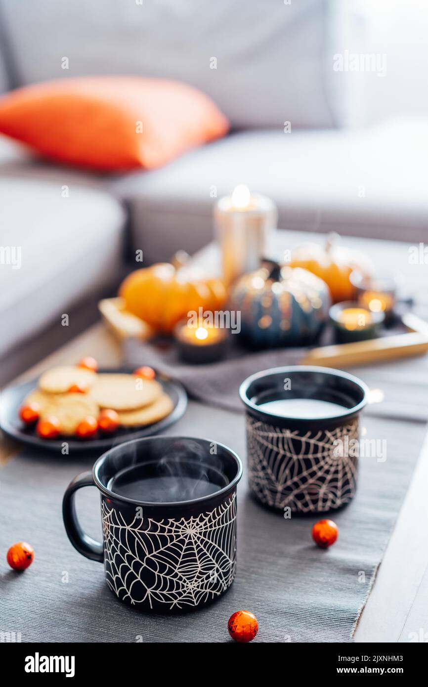 Accogliente Halloween piani a casa. Bevanda calda a base di tè in tazze nere con motivo a rete di ragno, biscotti e dolci sul piatto, decorazioni in zucca sul tavolino da caffè. Foto Stock