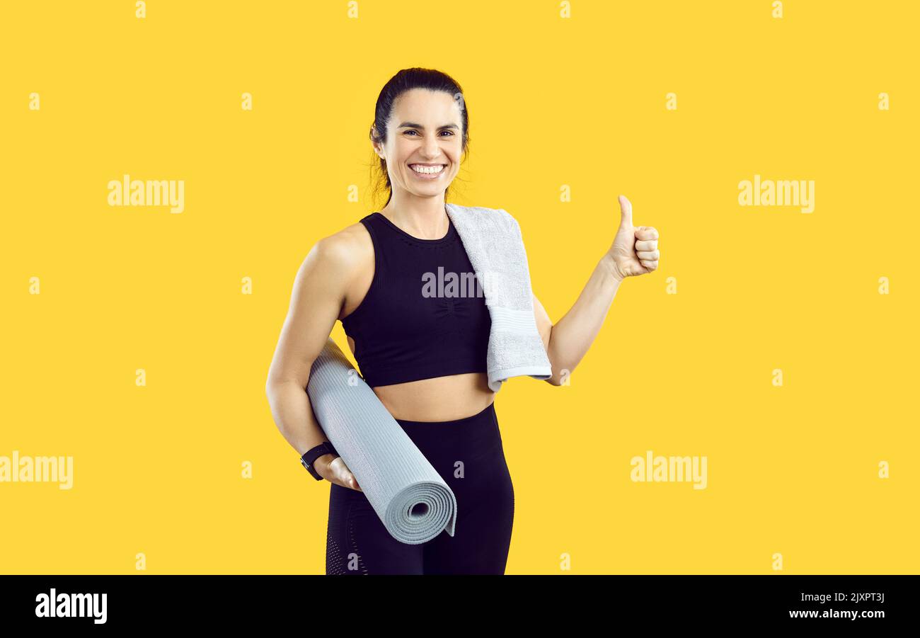 Studio shot di felice donna sportiva con asciugamano e tappetino da allenamento che dà i pollici e sorridere Foto Stock
