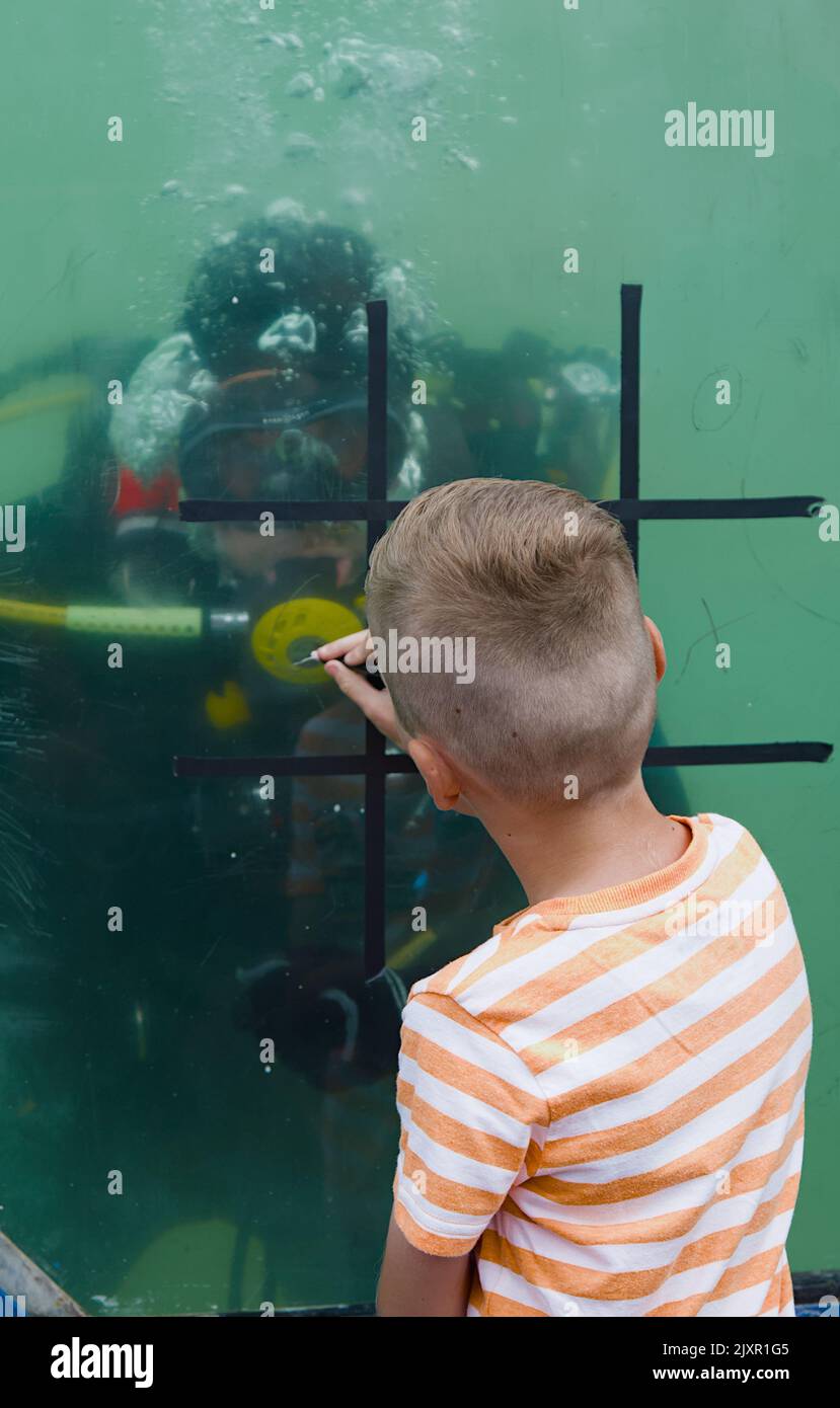 Young Boy Playing Noughts and Crocs, Tic TAC Toe Game Against A Royal Navy Diver wear Scuba Equipment in A Tank of Water, Bournemouth UK Foto Stock