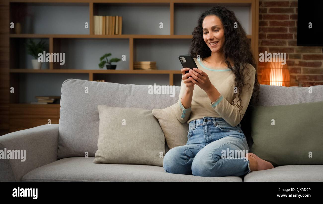 Sorridente Ispanica caucasica 30s donna casalinga ragazza seduta sul divano comodo divano a casa utilizzando smartphone per ordinare consegna di cibo chatting con Foto Stock