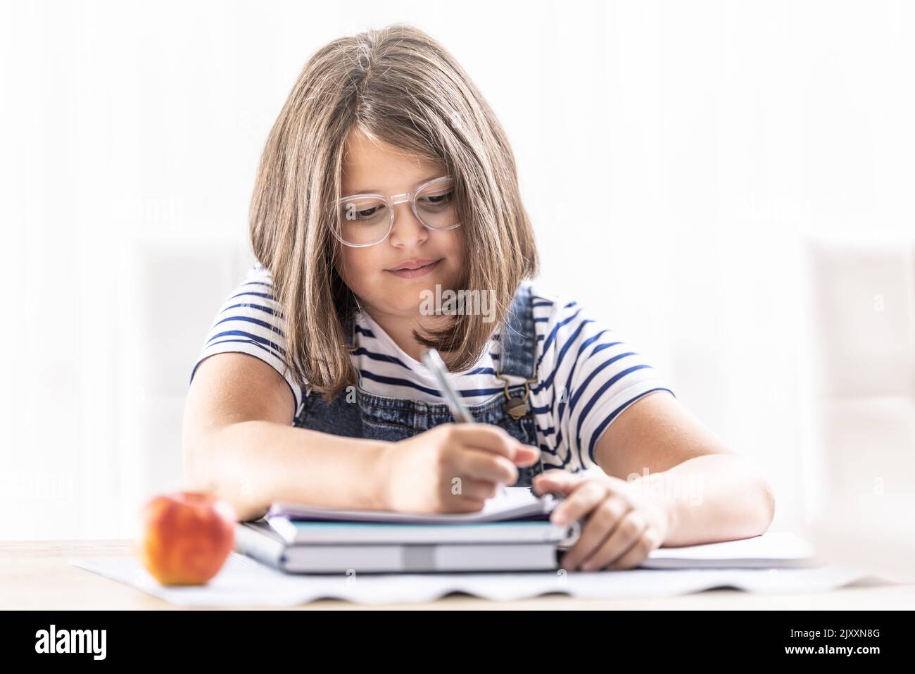 La scuola che frequenta la ragazza che indossa gli occhiali scrive in un libro di esercitazione con una penna che ha una mela su una tabella oltre lei. Foto Stock