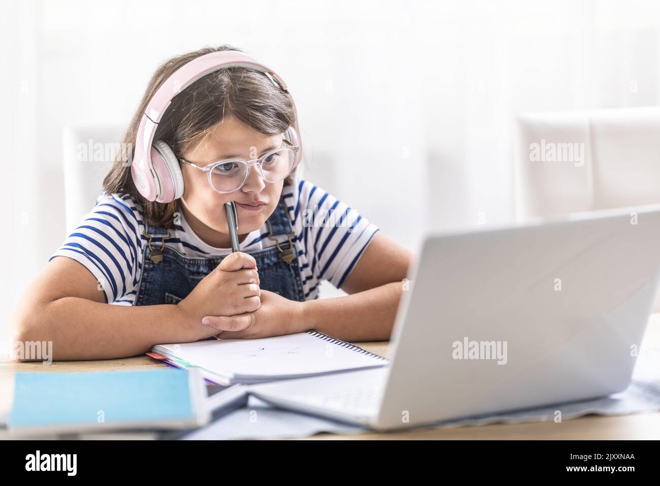 Ragazza in cuffie indossa occhiali e si appoggia contro la matita cercando di capire le cose che studia online. Foto Stock