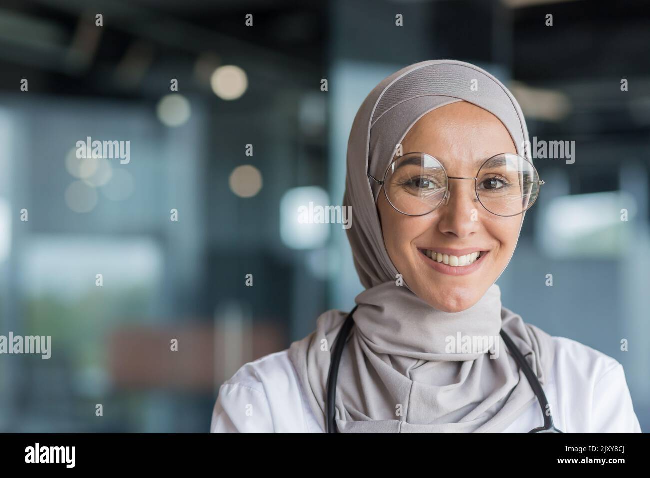 Primo piano ritratto fotografico di una dottoressa musulmana in hijab grigio, medico femminile sorridente e guardando la macchina fotografica, operatrice in occhiali e camice medico bianco e occhiali che lavorano all'interno di un moderno ufficio clinico Foto Stock