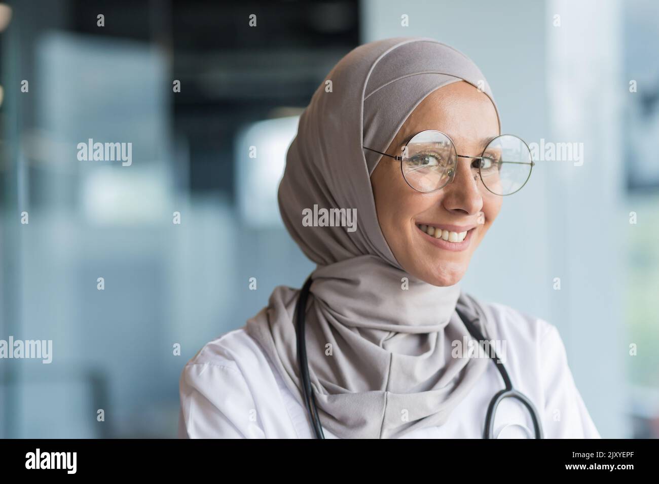 Primo piano ritratto fotografico di una dottoressa musulmana in hijab grigio, medico femminile sorridente e guardando la macchina fotografica, operatrice in occhiali e camice medico bianco e occhiali che lavorano all'interno di un moderno ufficio clinico Foto Stock