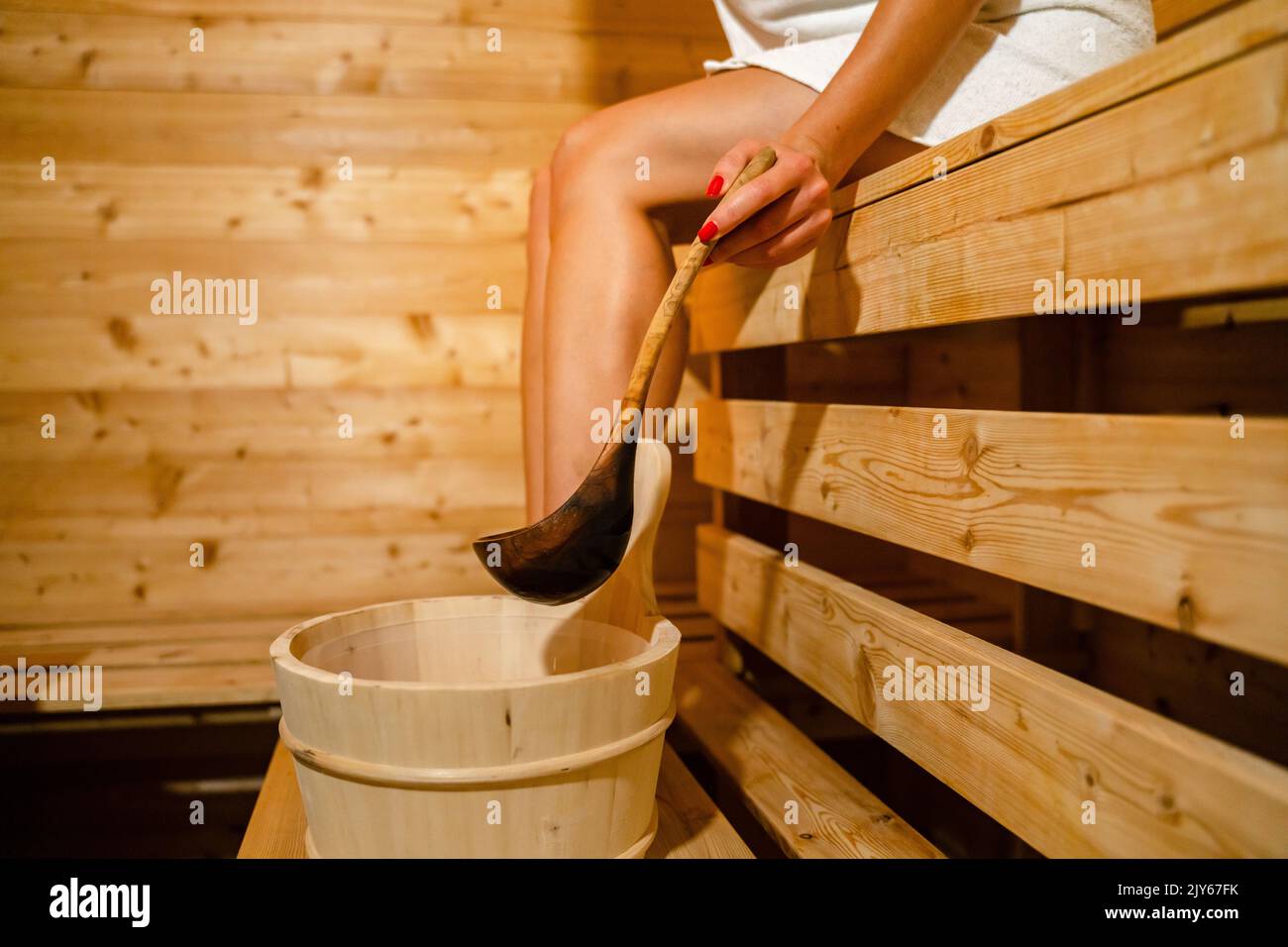 mano di donna caucasica sconosciuta in sauna spa prendere l'acqua dal secchio con cucchiaio di legno per versare sulle pietre calde Foto Stock