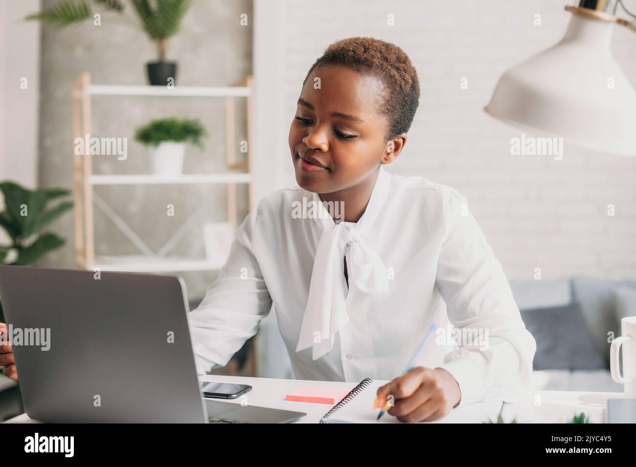 Foto di allegra donna da corsa mista in camicia bianca che lavora con il portatile mentre si siede al tavolo in ufficio. Lavoro di squadra, meeting di lavoro. Freelance Foto Stock