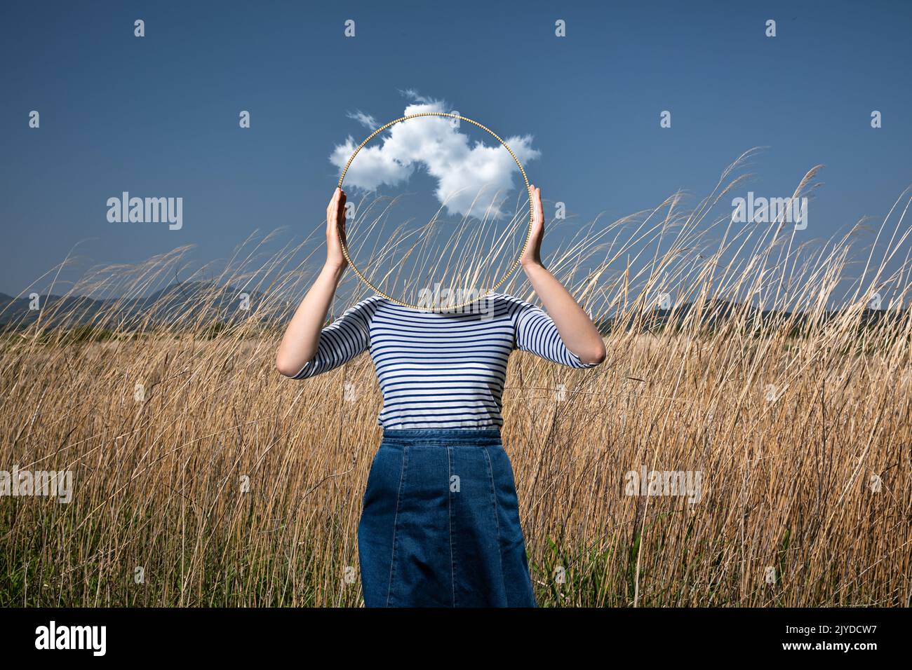 Surrealismo con una donna che tiene uno specchio e copre il suo volto nel campo con uno sfondo trasparente dietro lo specchio Foto Stock