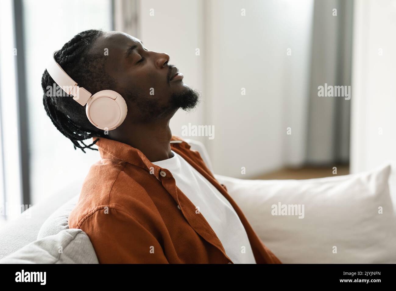 Uomo nero rilassato in cuffie seduto sul divano con gli occhi chiusi, ascoltando musica a casa, vista laterale, spazio copia Foto Stock
