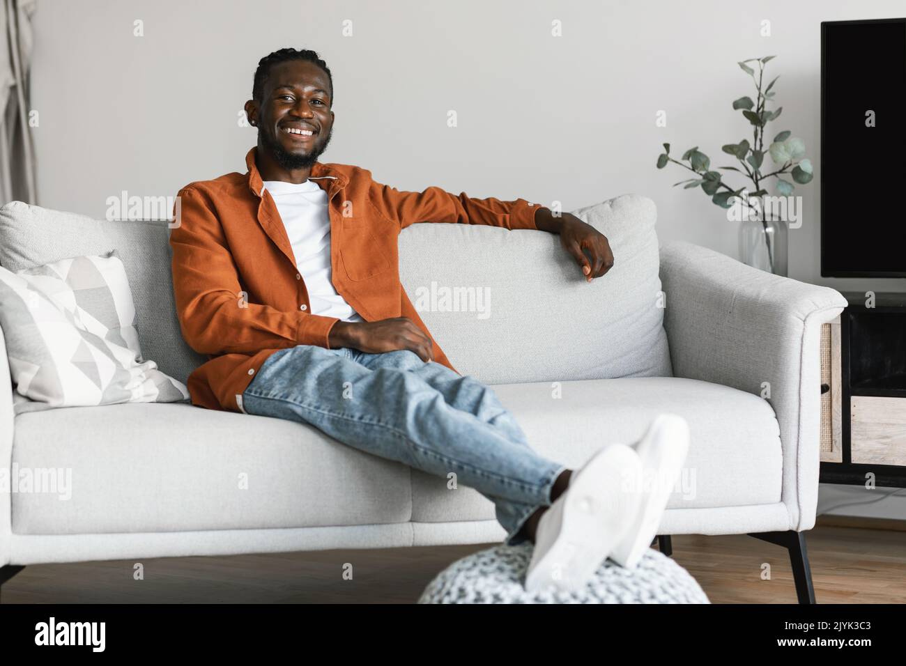 Rimani a casa da solo, goditi il silenzio. Felice uomo afroamericano seduto sul divano in un accogliente soggiorno luminoso, spazio copia Foto Stock