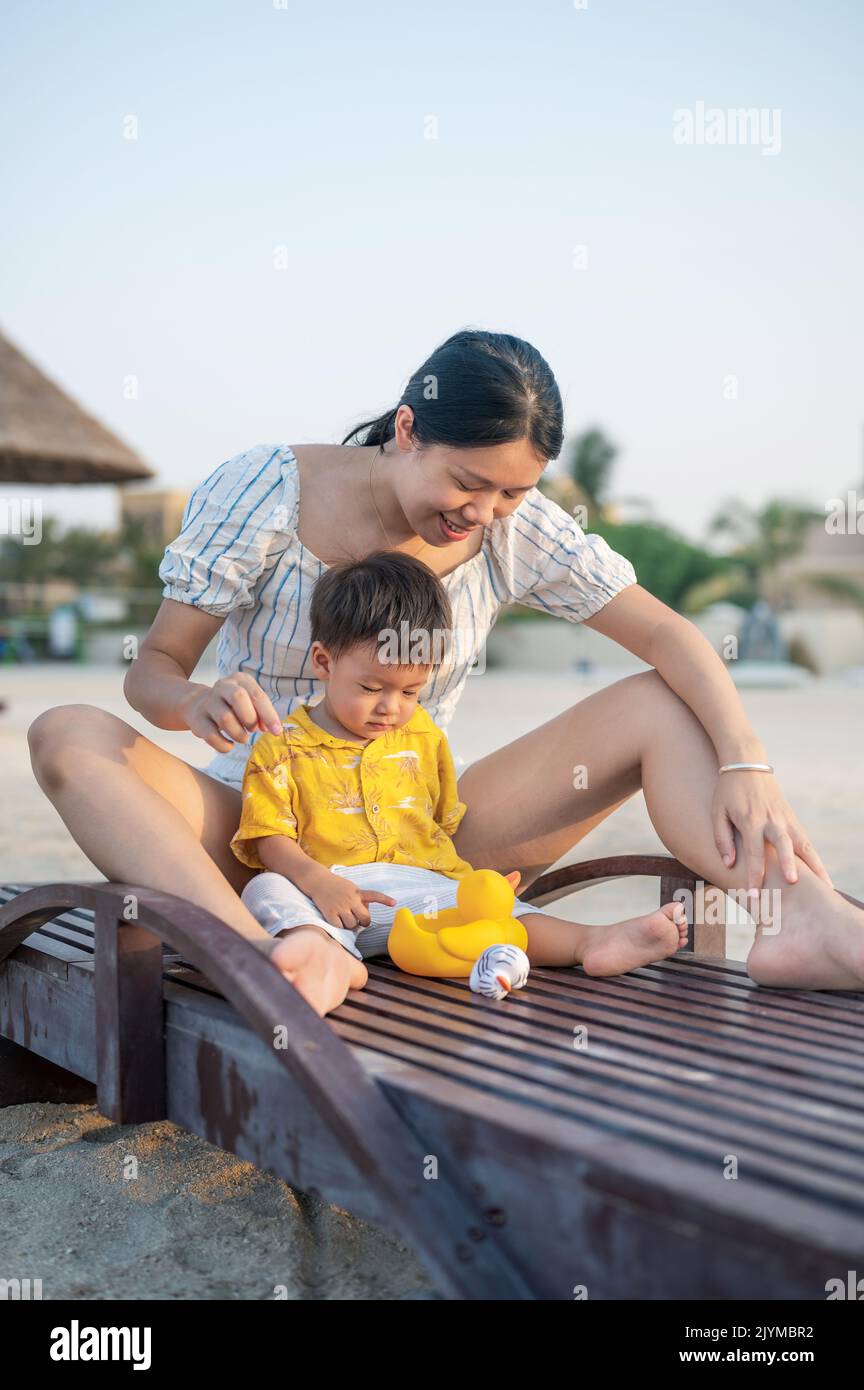 Bambino in vacanza sulla spiaggia seduto sul lettino con la madre e giocando con l'anatra giocattolo. Donna asiatica e il suo bambino di un anno che si divertono Foto Stock