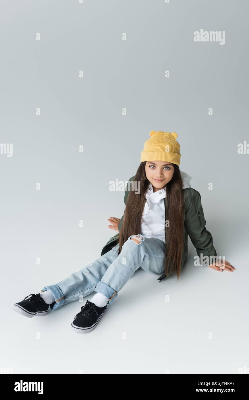 vista ad angolo alto di adorabile ragazza in abito autunnale alla moda e berretto beanie seduto sul grigio, immagine stock Foto Stock