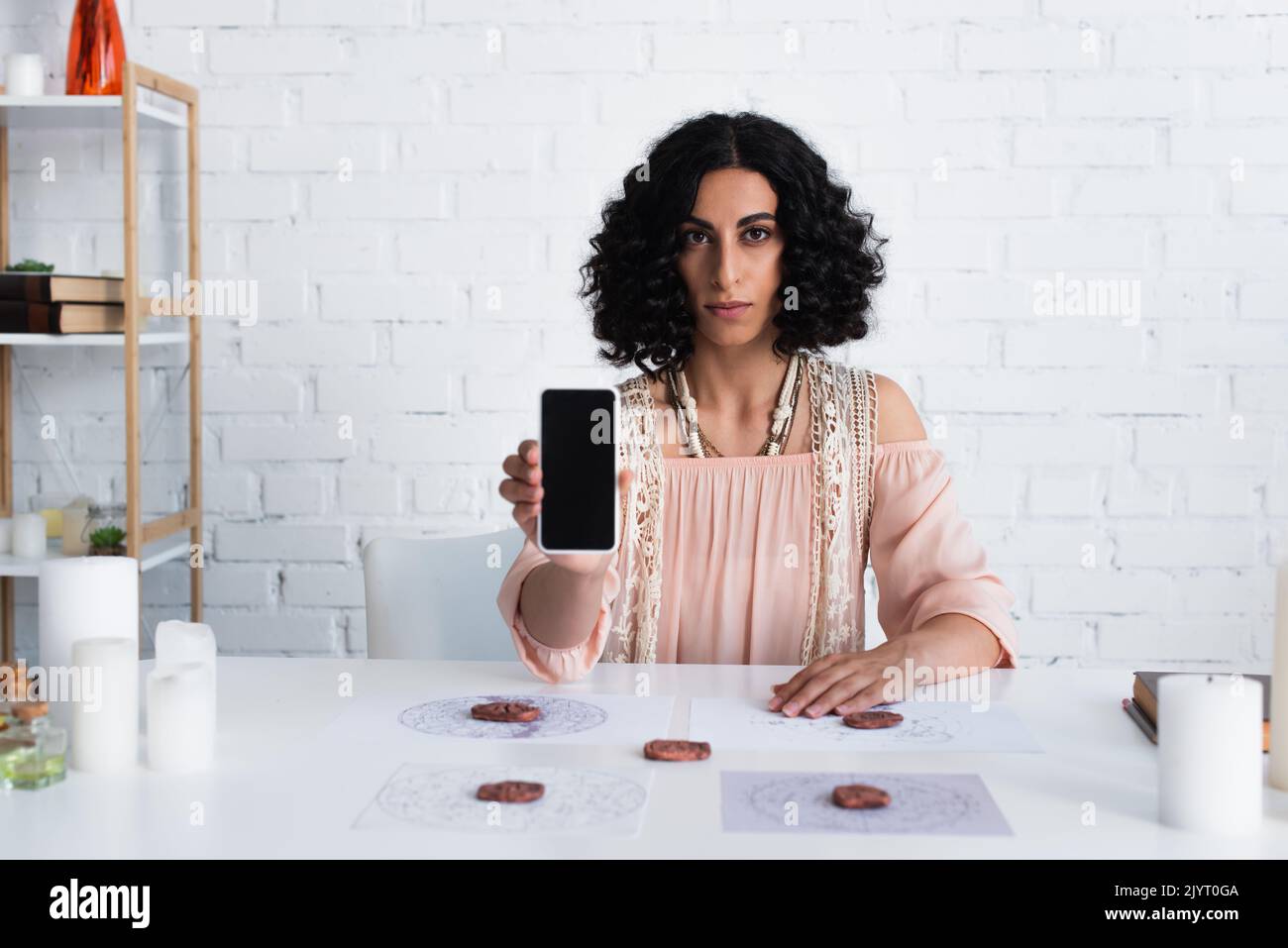 bruna fortuna che guarda la fotocamera mentre tieni lo smartphone con schermo vuoto vicino a rune di argilla e mappe delle stelle Foto Stock