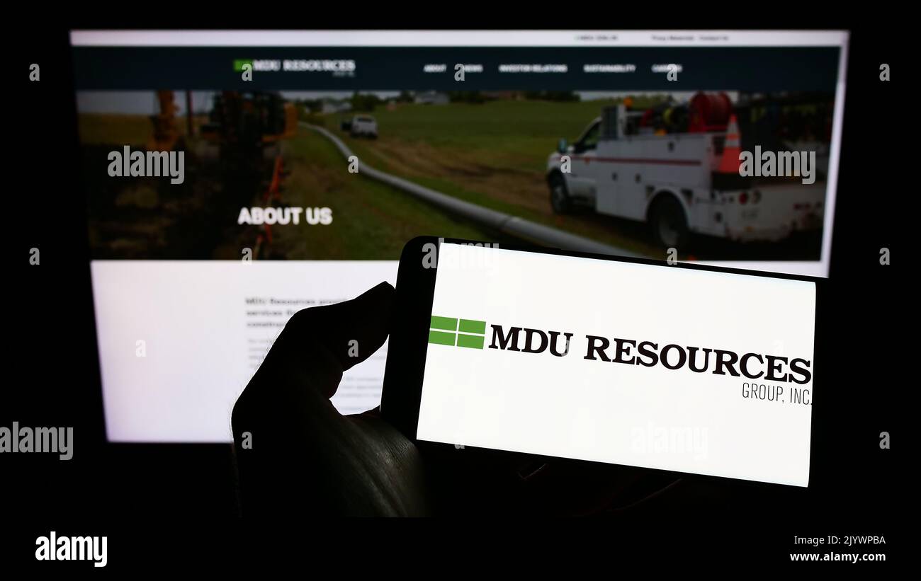 Persona che tiene il cellulare con il logo della società americana MDU Resources Group Inc. Sullo schermo di fronte alla pagina web aziendale. Messa a fuoco sul display del telefono. Foto Stock