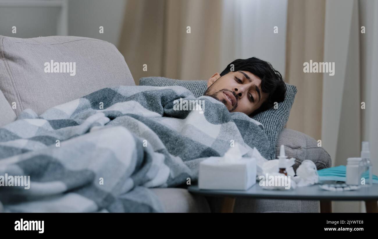 Indiano sopportato triste uomo esausto sdraiato sul divano di casa coperta malata che soffre di febbre sintomi malattia coronavirus concetto guardando Foto Stock