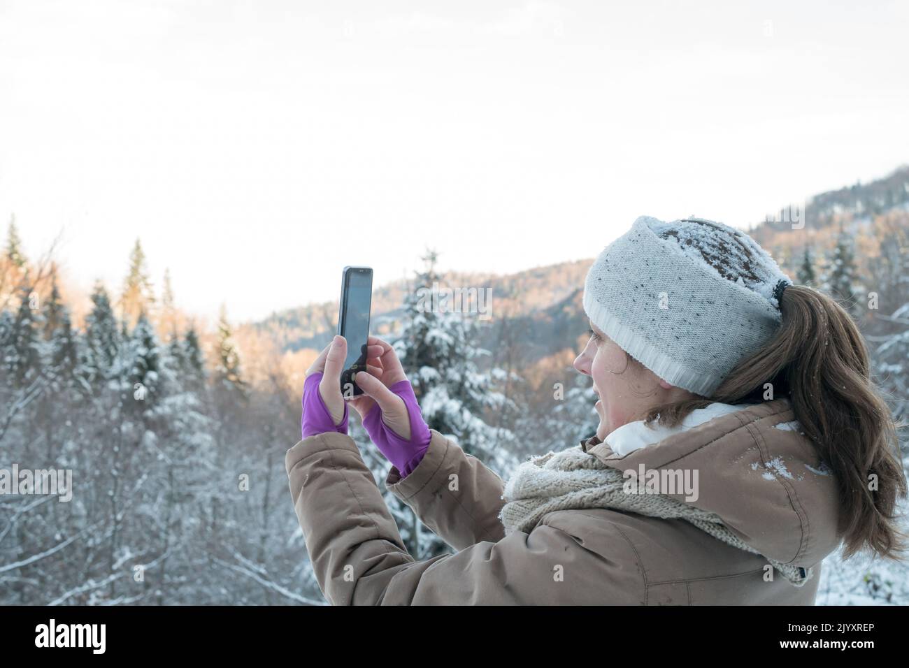 Donna che guarda al display mobile, fotografa i paesaggi invernali della natura. Ragazza caucasica di mezza età con telefono cellulare nella foresta coperta di neve. Foto Stock
