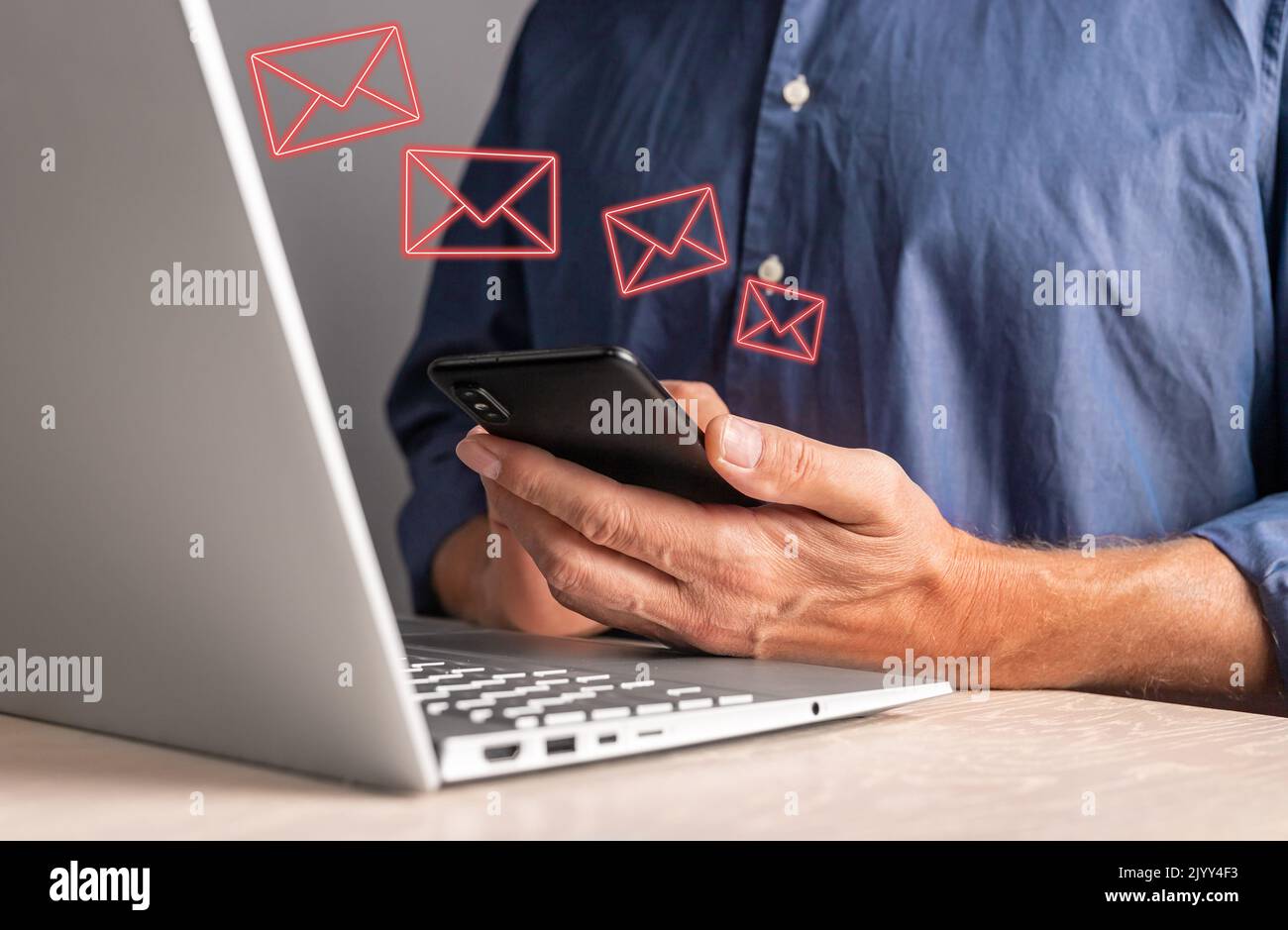 Uomo che riceve e-mail di phishing al telefono mentre lavora sul computer portatile. Maschio seduto al tavolo con il computer e tenendo lo smartphone. Messaggi indesiderati, spam te Foto Stock