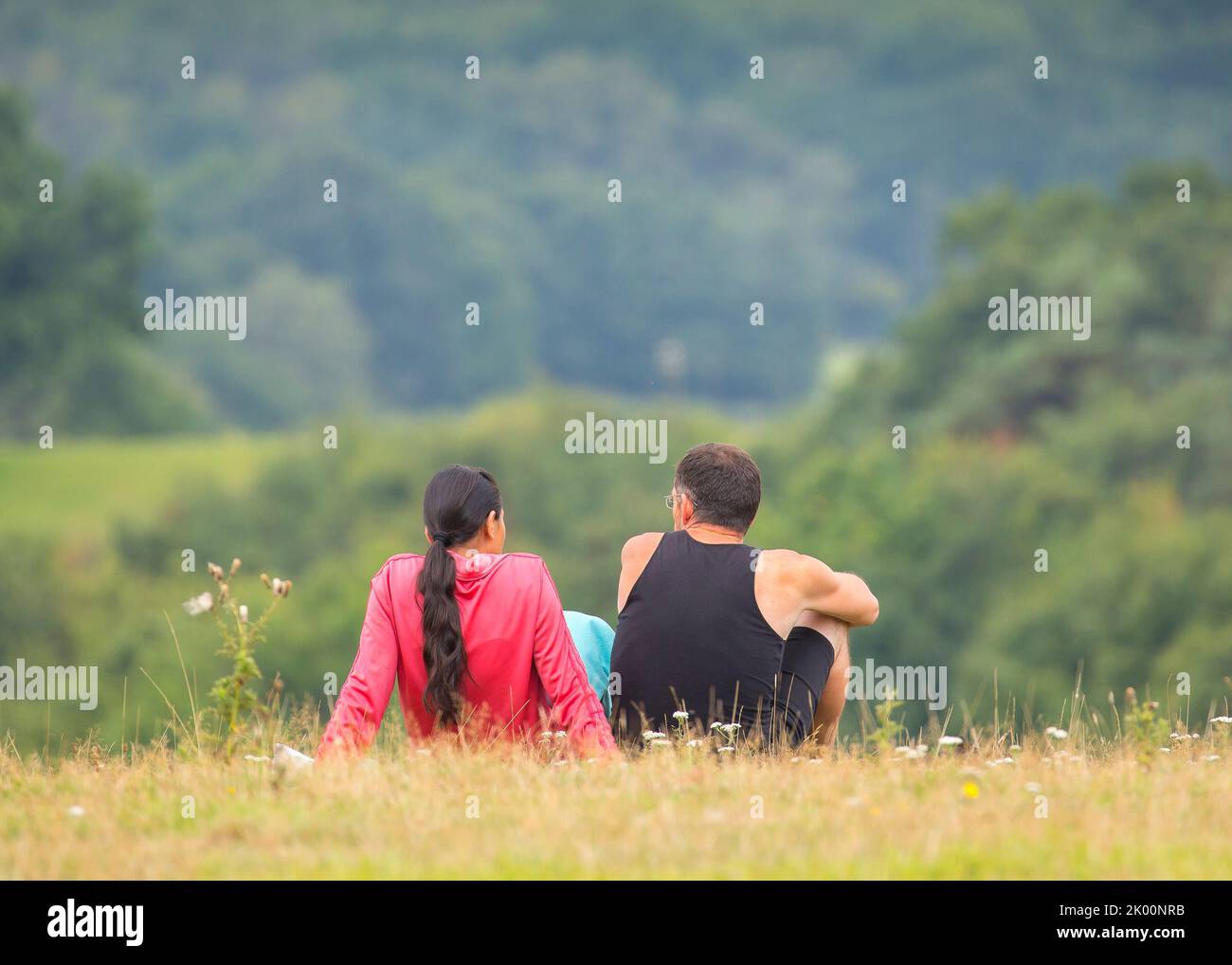 Vista posteriore/posteriore di un uomo e di una donna seduti a rilassarsi insieme e fare una pausa dalla loro passeggiata su una collina rurale del Regno Unito, in estate, guardando la vista. Foto Stock