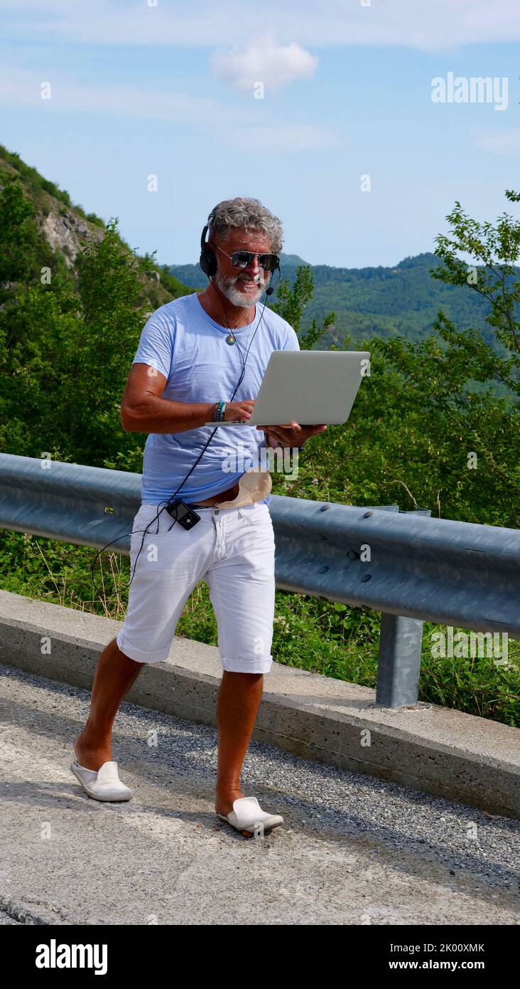 Uomo anziano dai capelli grigi diabetico malato freelancer con barba in occhiali da sole, lavorando su un computer portatile sulla natura, montagne. Un vecchio pazzo e straordinario che cammina per strada in estate. Verticale Foto Stock