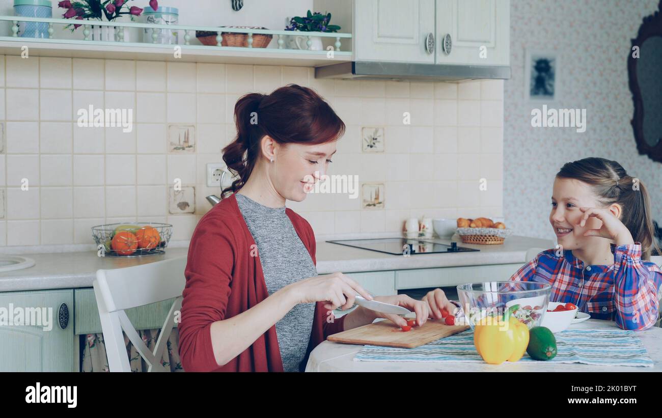 Allegra madre felice che taglia i pomodori mentre cucinava e che parla con la sua figlia sorridente che alimenta la mamma in cucina a casa. Concetto di famiglia, cuoco e gente Foto Stock