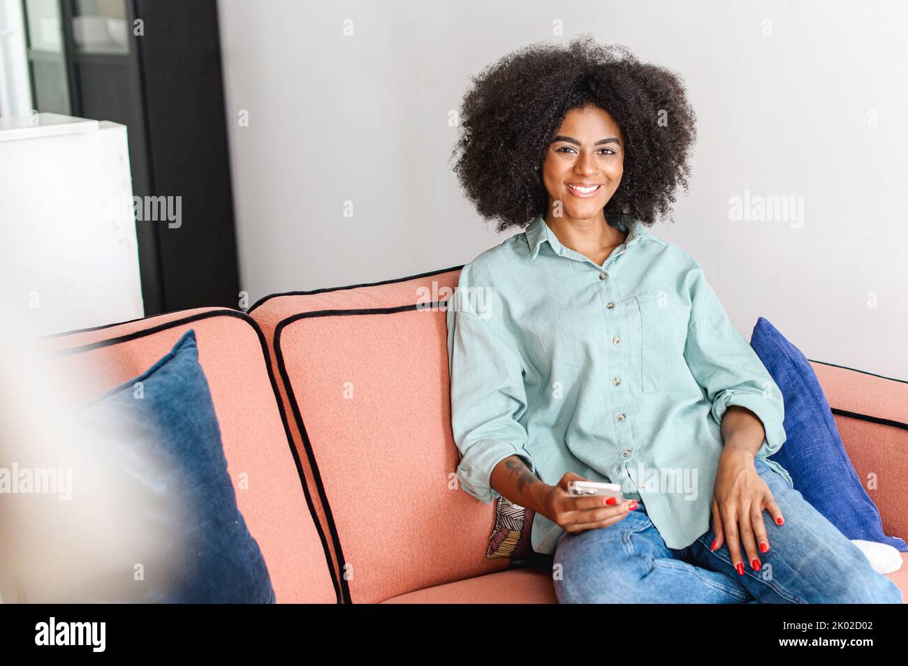 Ritratto di bella donna nera con capelli ricci naturali afro guardando la fotocamera in un'atmosfera accogliente casa, affascinante ragazza serena e spensierata tenendo smartphone e riposo a casa Foto Stock