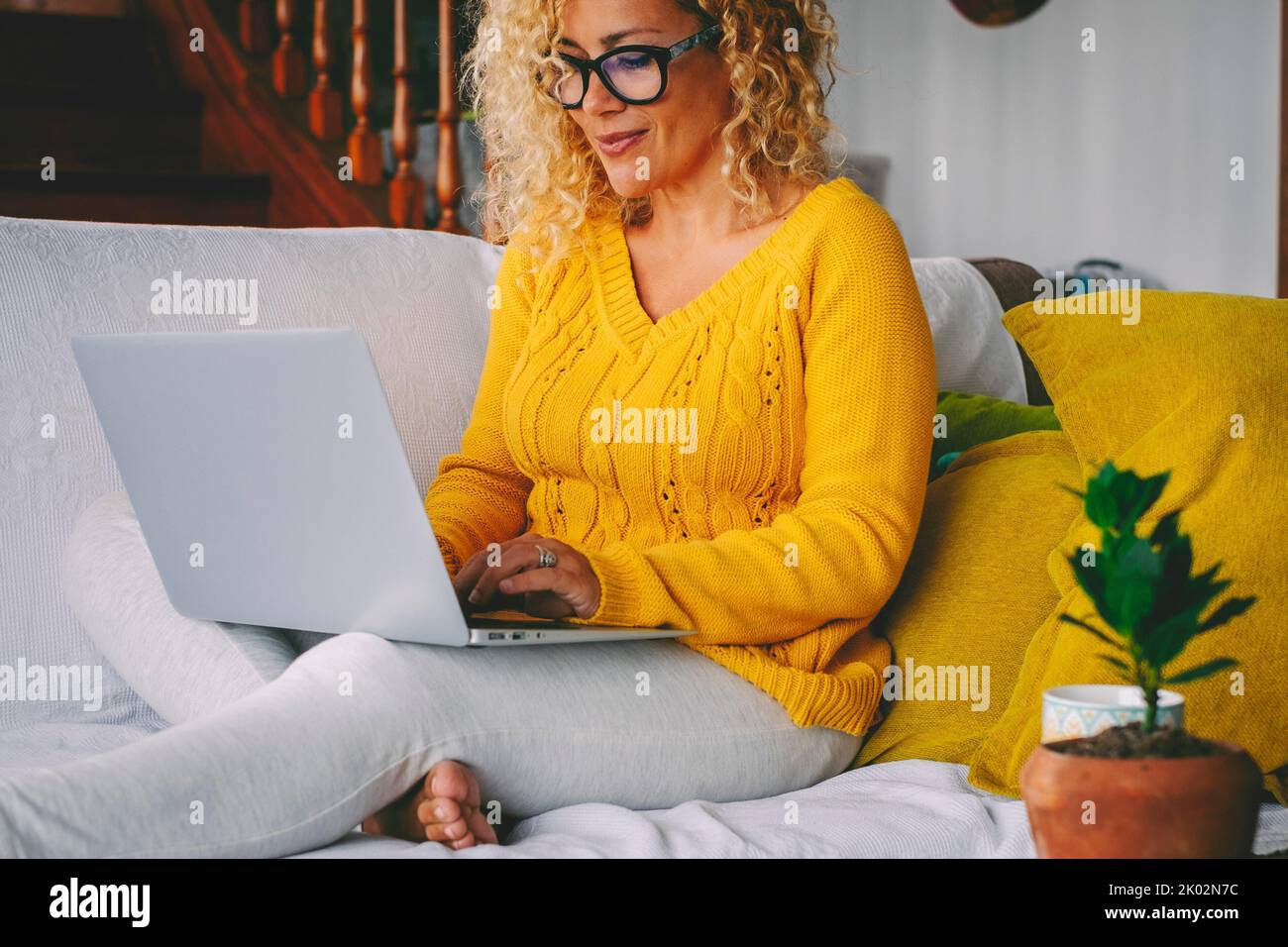 Donna piuttosto di mezza età che scrive sul laptop e sorridente. Una donna che usa il computer da sola a casa. Donna bionda adulta godere del tempo libero con connessione wireless notebook seduta e rilassante Foto Stock