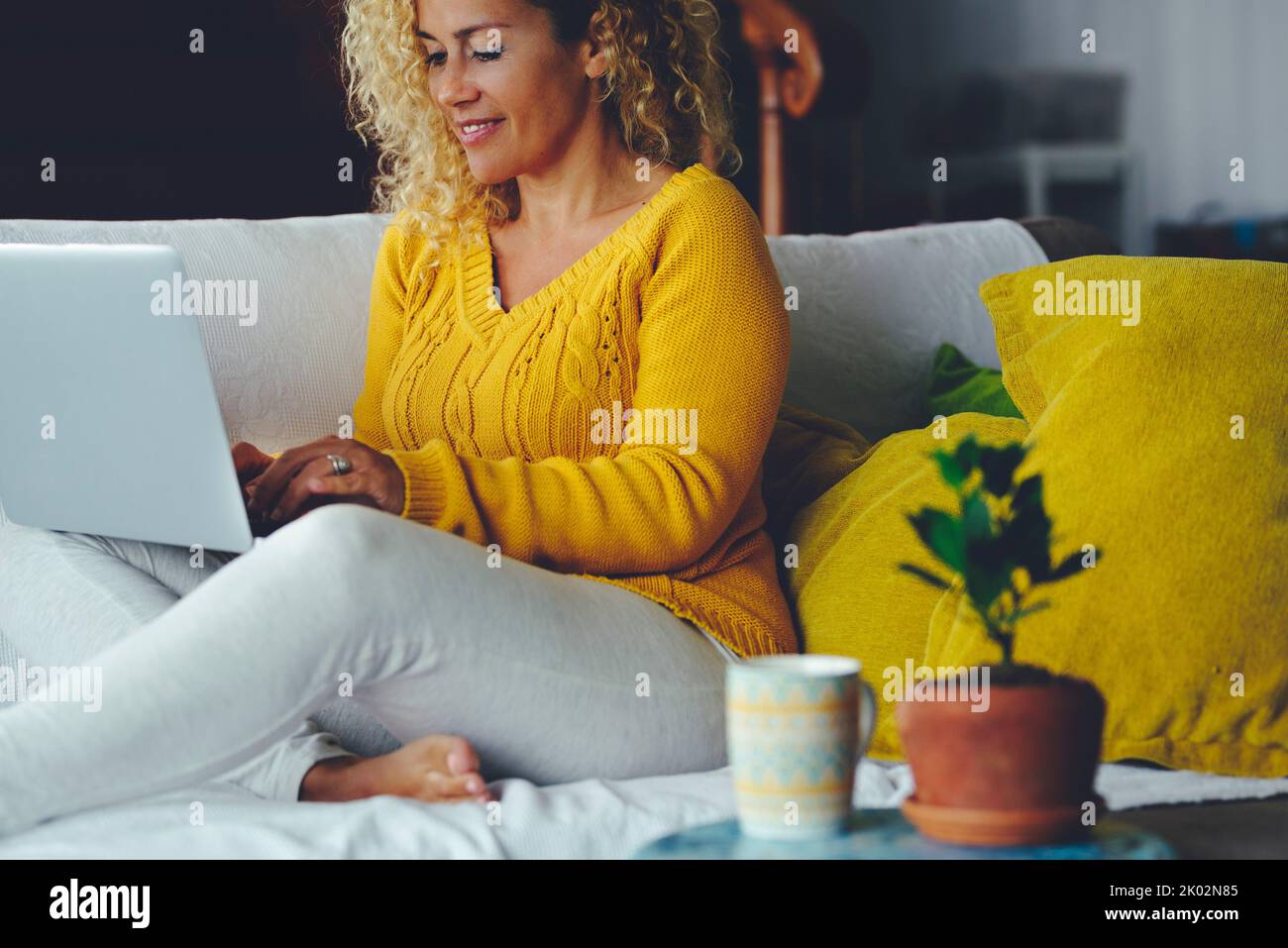 Una donna a casa seduta sul divano navigando sul web e utilizzando il laptop per attività di connessione tecnologica. Le donne moderne scrivono sul computer da sole comodamente sul divano e sorridono Foto Stock