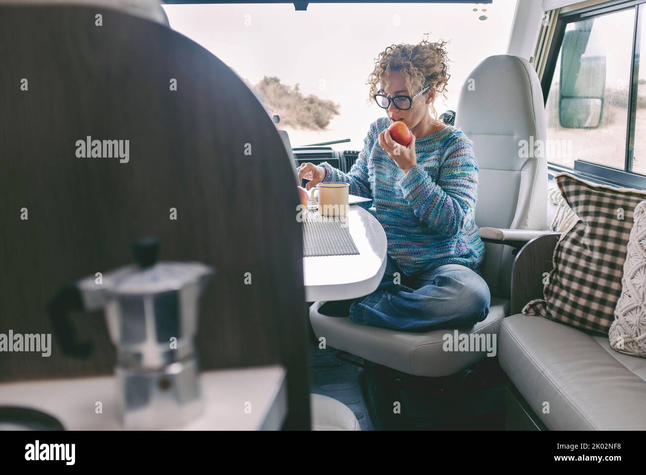Vista della giovane donna seduta e che ha tempo libero all'interno di un camper casa a motore. Stile di vita sano femmina mangiare mela e utilizzare computer portatile. Stile di vita alternativo gratuito fuori rete Foto Stock