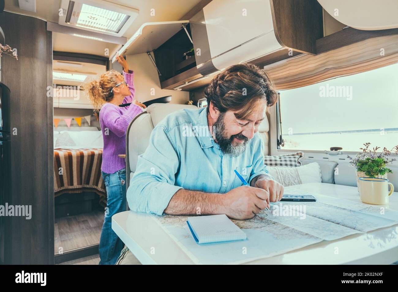 Barbuto uomo che progetta la prossima destinazione vacanza di viaggio seduto al tavolo all'interno del suo camper van. Donna in background cucinare e gestire lo stoccaggio. Vacanza vita vacanza Foto Stock