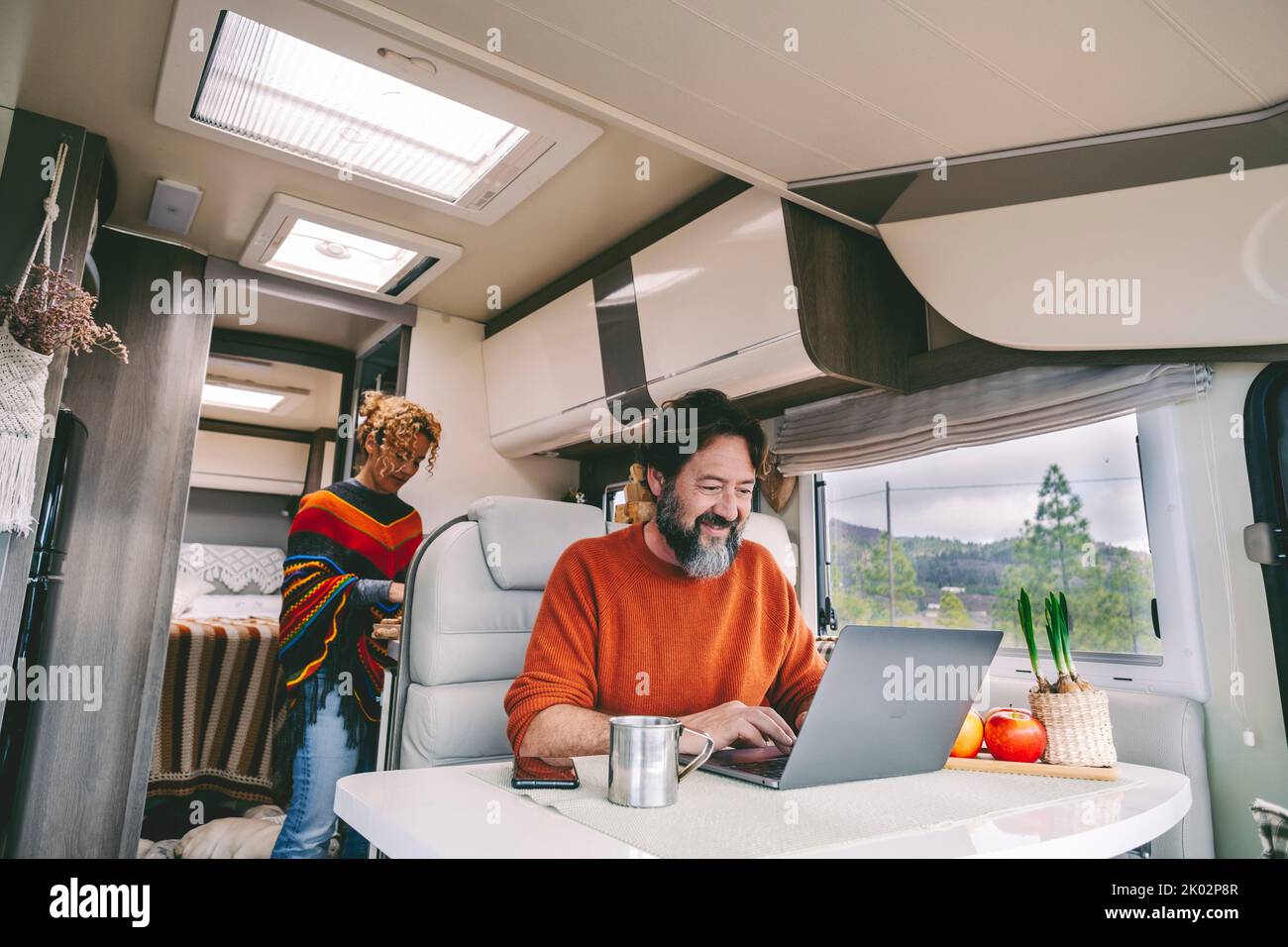 Vita normale quotidiana all'interno di un camper camper casa mobile rv. Donna che cucinava in cucina e uomo adulto che lavorava sul computer portatile. All'aperto parco natura fuori dalla finestra. Libertà vanlife persone lifestyle Foto Stock