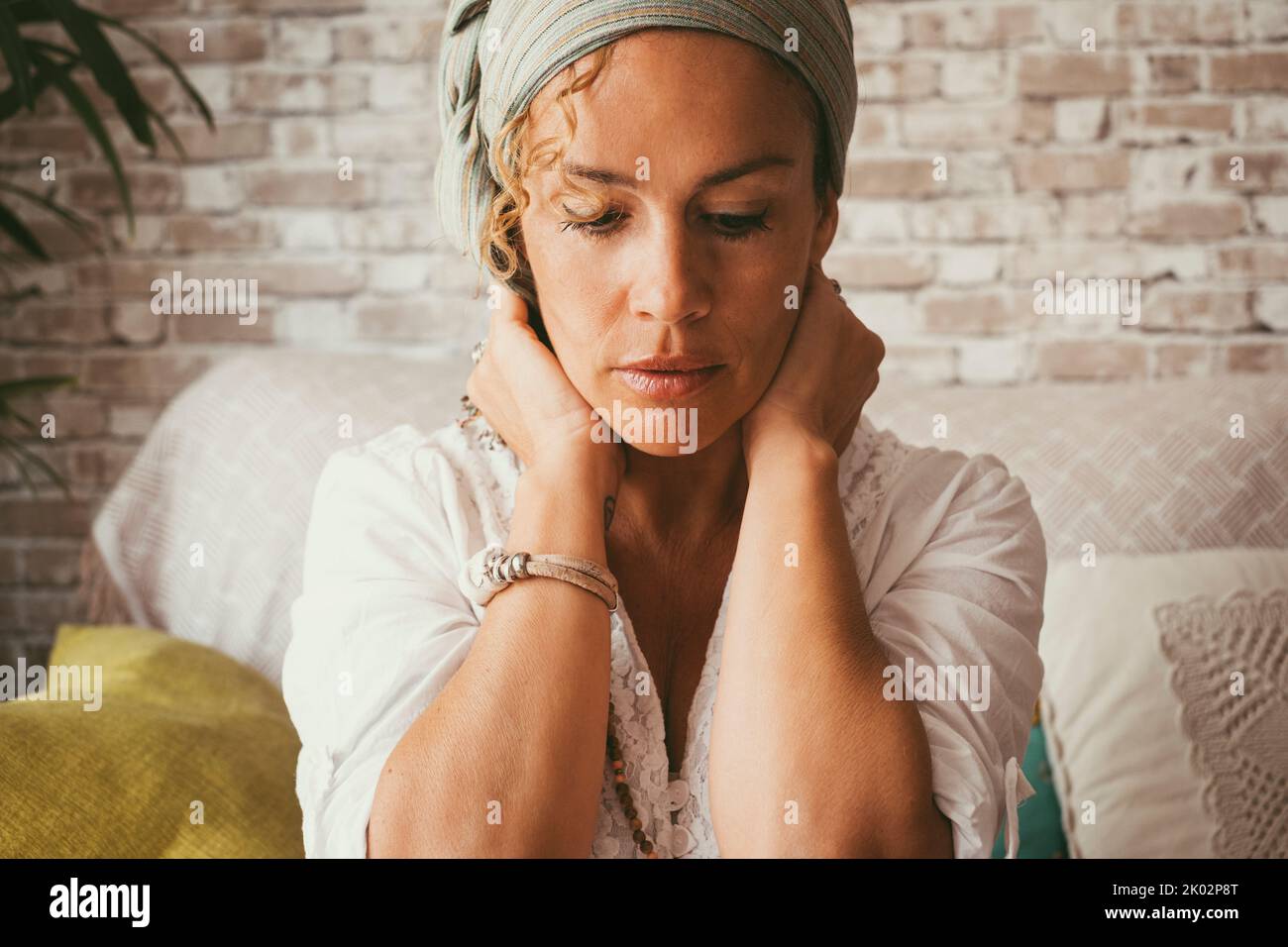 Gente adulta donna che tocca il collo con l'espressione preoccupata sul viso. Concetto di problemi di vita e di donna. Solitaria donna che soffre depressione e solitudine solo a casa seduta sul divano guardando verso il basso Foto Stock