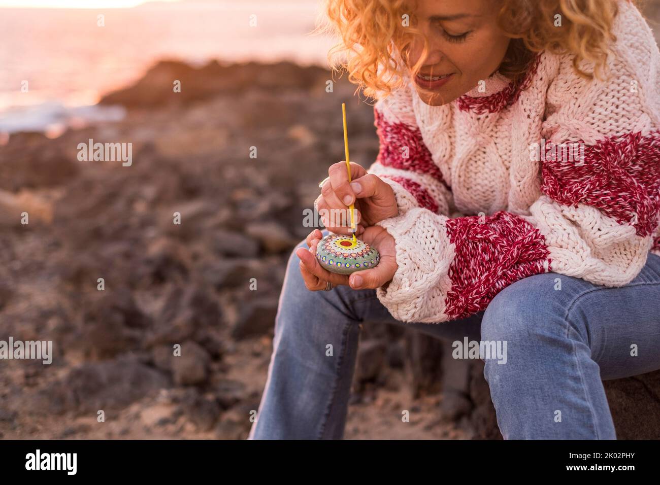 Donna adulta dipingere una pietra con mandala design all'aperto in attività ricreative artistiche. Felice femmina godere pennello e colori e creare arte su pietre. Luce del tramonto sullo sfondo Foto Stock