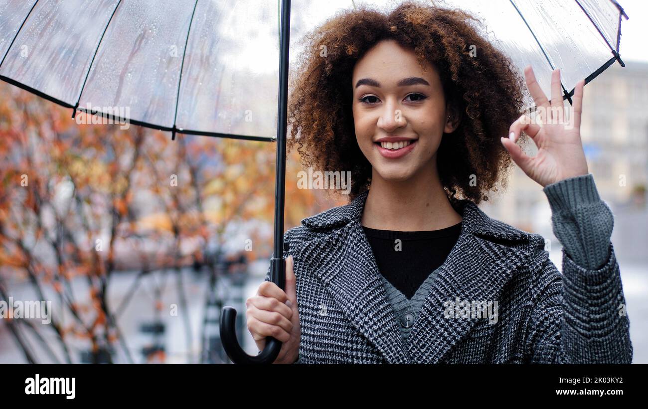 Bella donna felice di successo riccia ragazza afroamericana con ombrello trasparente stand in autunno città sotto la pioggia guardando la macchina fotografica sorridente Foto Stock