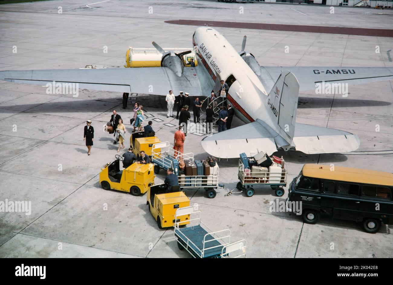 A Dan Air Douglas DC-3 Dakota, C-47, registrazione G-AMSU all'aeroporto di Manchester in Inghilterra il 28th giugno 1964. I passeggeri sono visti sbarcare dall'aereo con vari veicoli aeroportuali intorno. Tipico dei viaggi aerei negli anni '1960s. Foto Stock