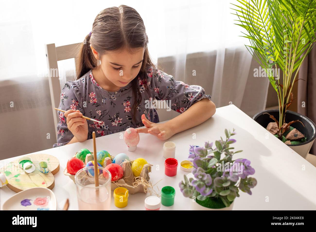 Una adorabile bambina siede su un tavolo bianco vicino alla finestra, si prepara per la festa di Pasqua Foto Stock