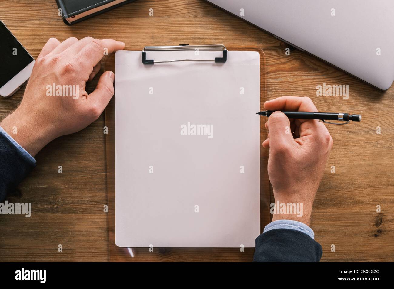 Business mockup, layout. Vista dall'alto della clipboard con foglio bianco di carta disposto verticalmente su un tavolo di legno. Le mani maschili tengono la matita o la punta a sfera Foto Stock