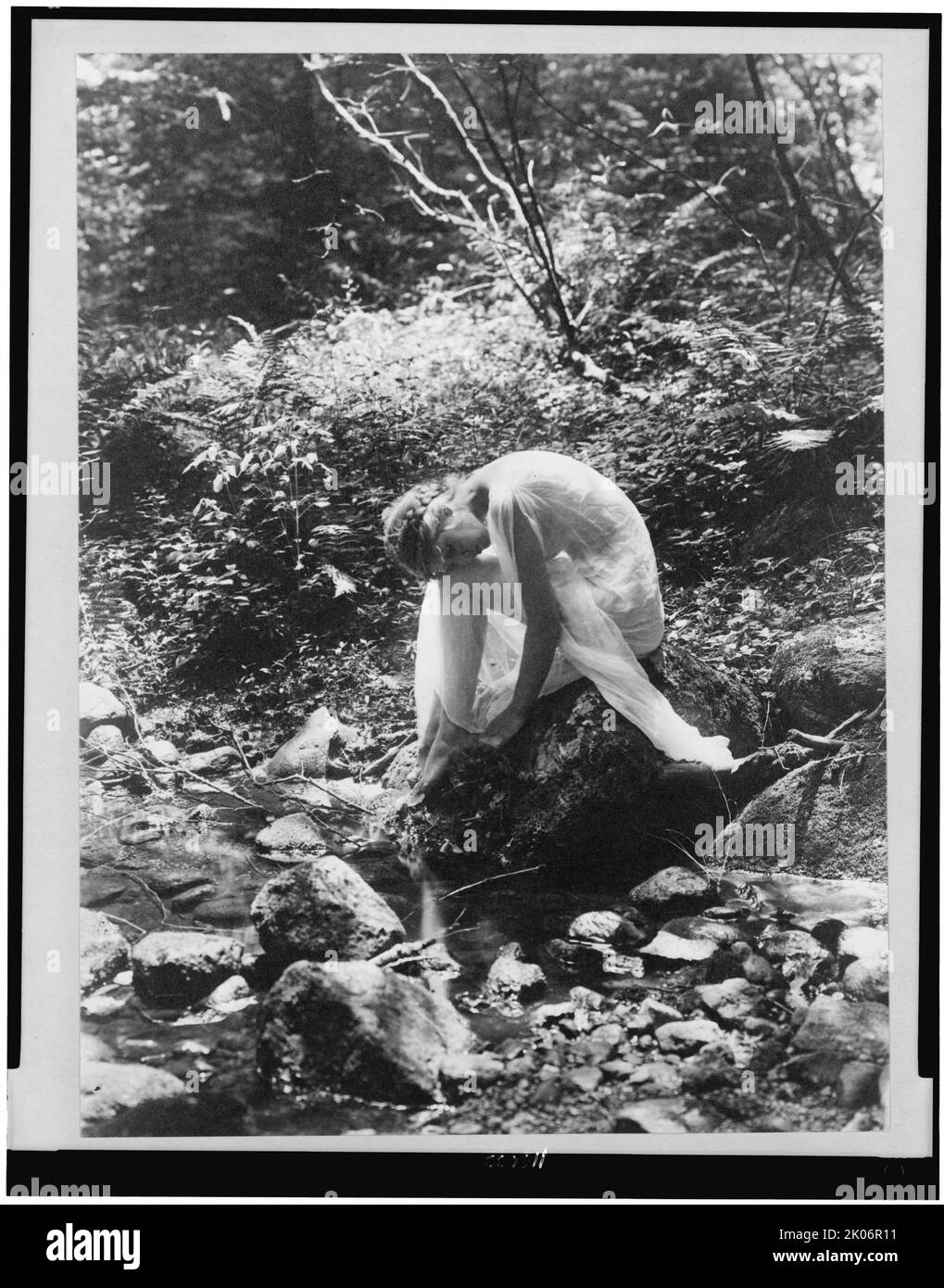 Legno-ninfa, tra 1900 e 1915. Donna in drappeggi classici seduti sulla roccia vicino al ruscello nei boschi. Foto Stock