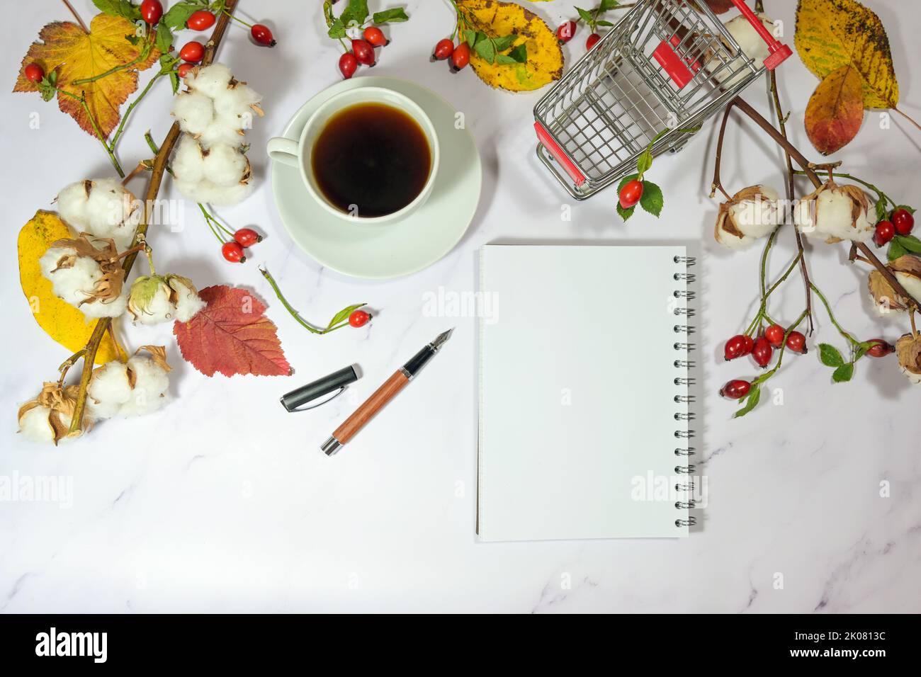 taccuino vuoto mock up, carrello per la spesa e caffè tazza su un desktop di marmo chiaro con colorate foglie autunnali, fianchi di rosa e rami di cotone, festa sal Foto Stock
