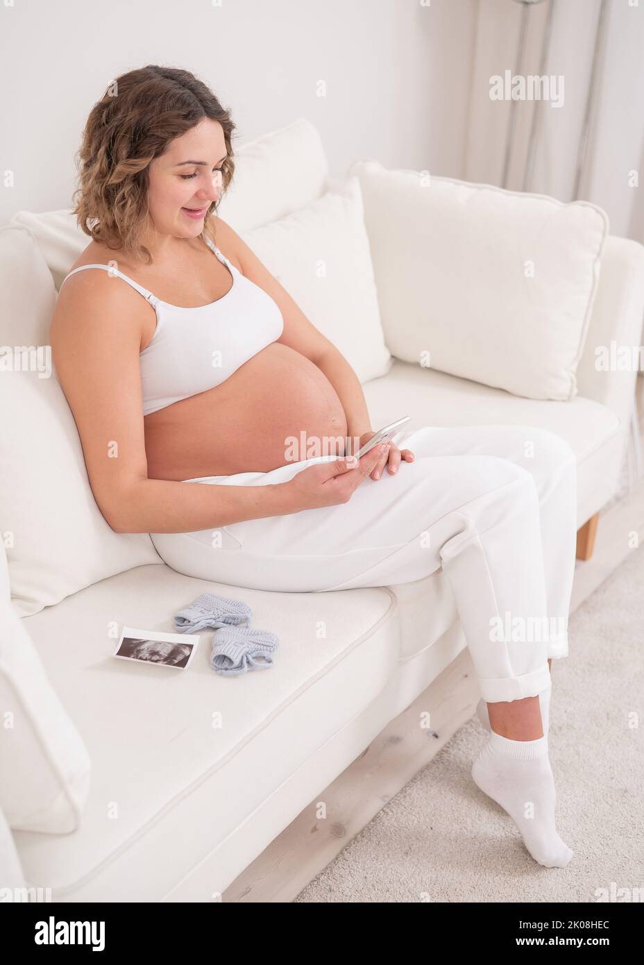 Una donna incinta siede su un divano bianco e tiene uno smartphone. Screening fetale a ultrasuoni. Foto Stock