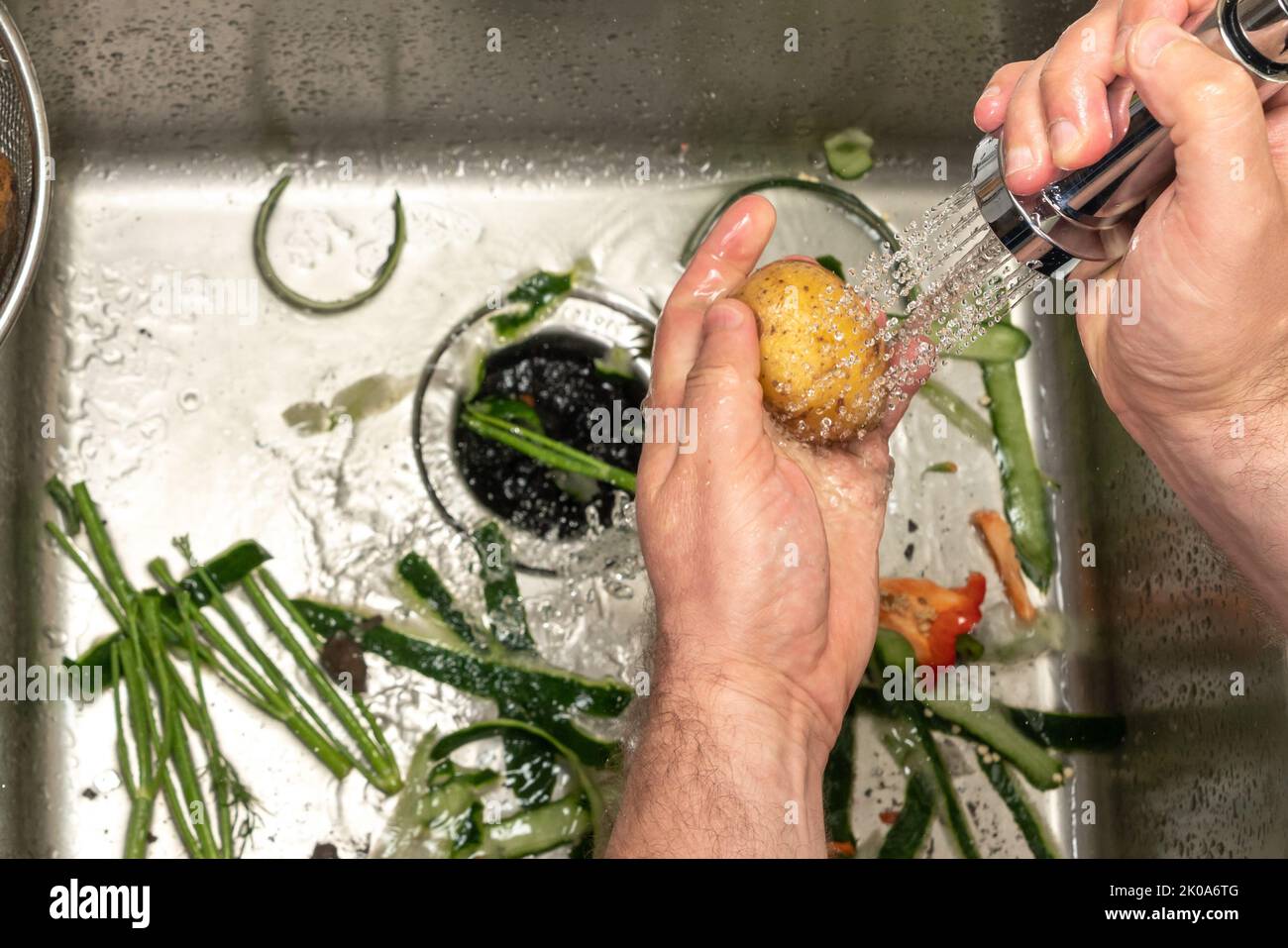 Un uomo lava le patate su un lavello da cucina riempito di rifiuti da altre verdure. Foto Stock