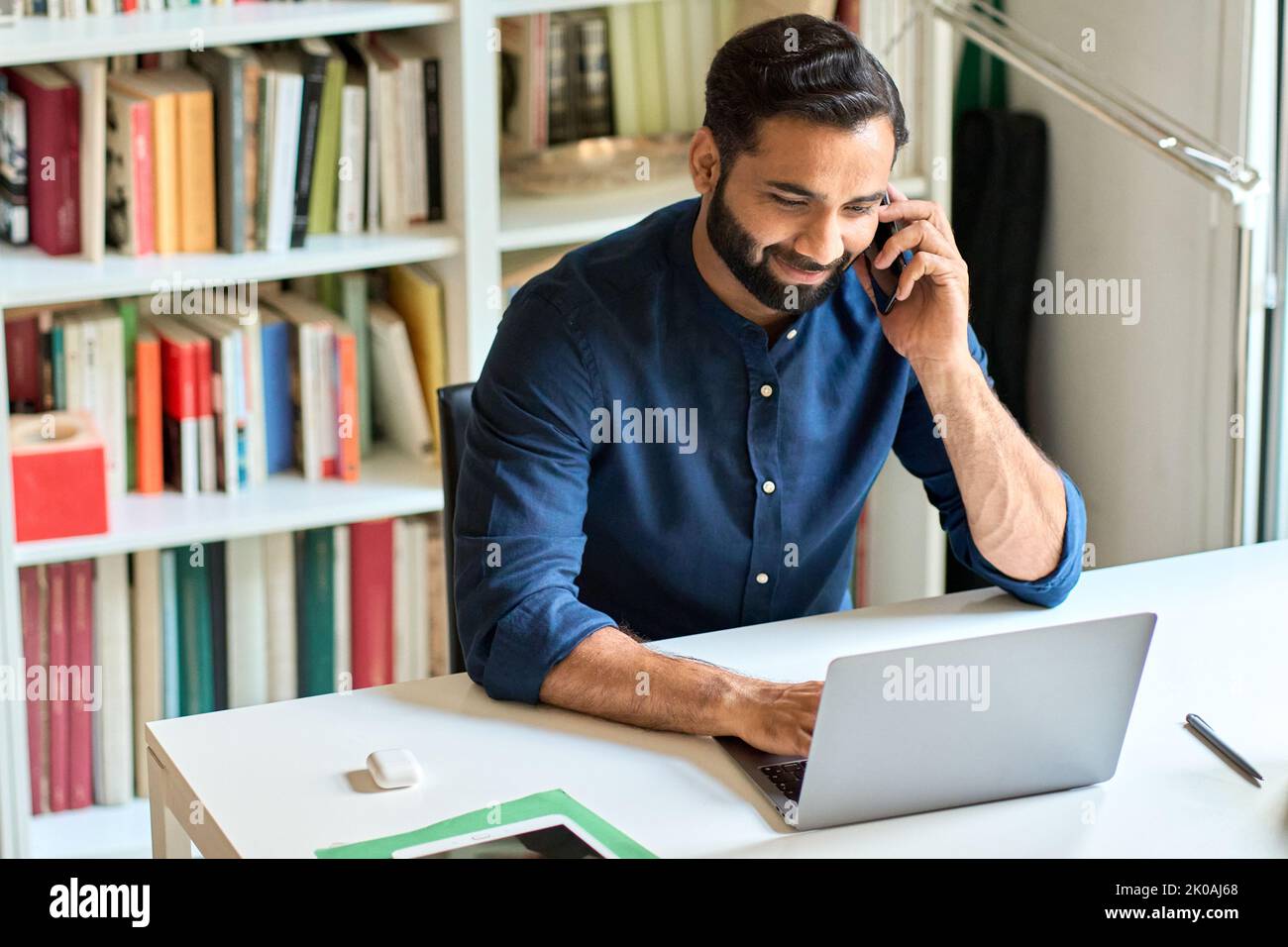 Uomo d'affari indiano sorridente seduto alla scrivania che parla al telefono che effettua una chiamata. Foto Stock