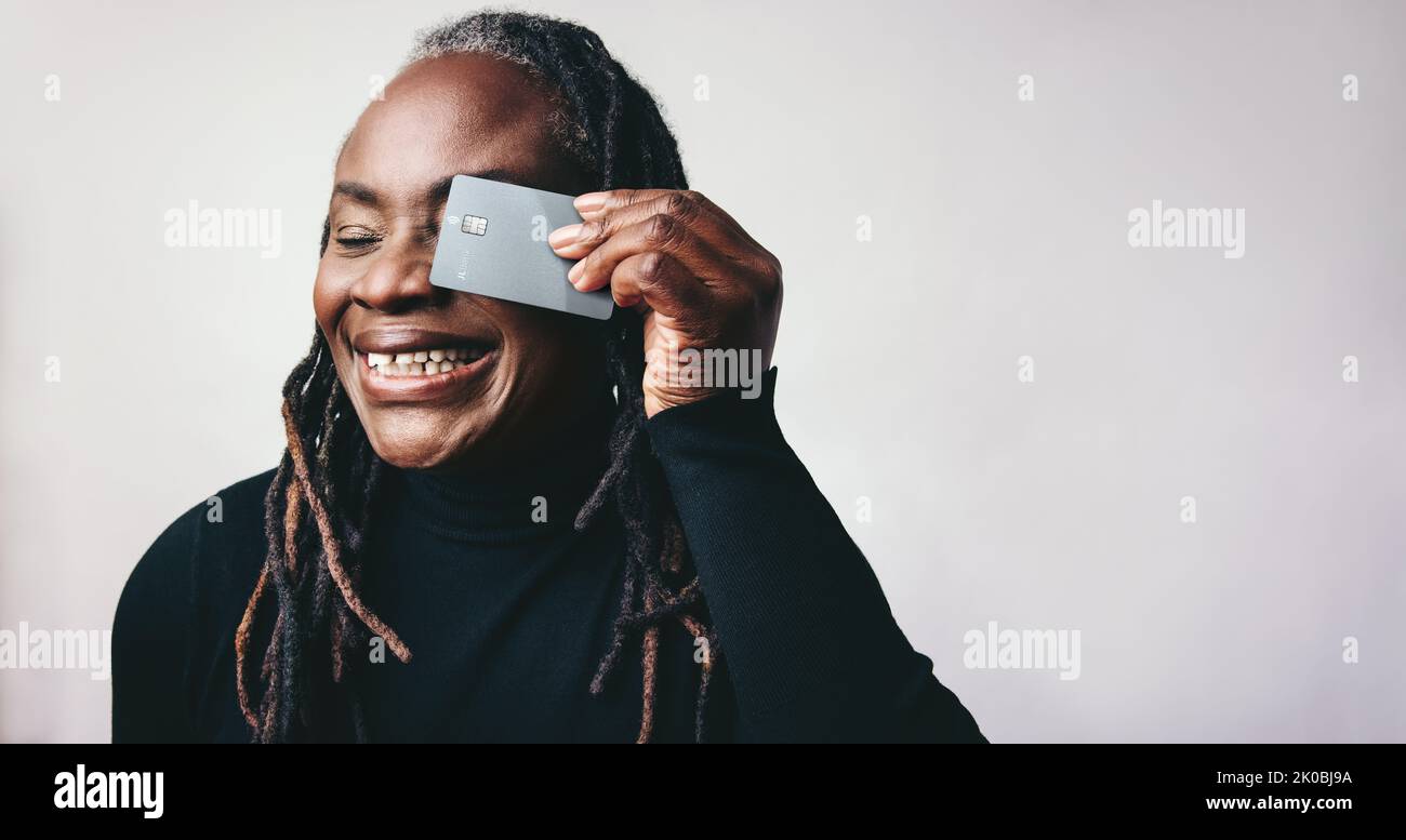 Donna matura allegra con greggi sorridenti mentre tiene una carta di credito davanti al suo occhio. Donna felice di mezza età che consiglia il sistema bancario elettronico Foto Stock