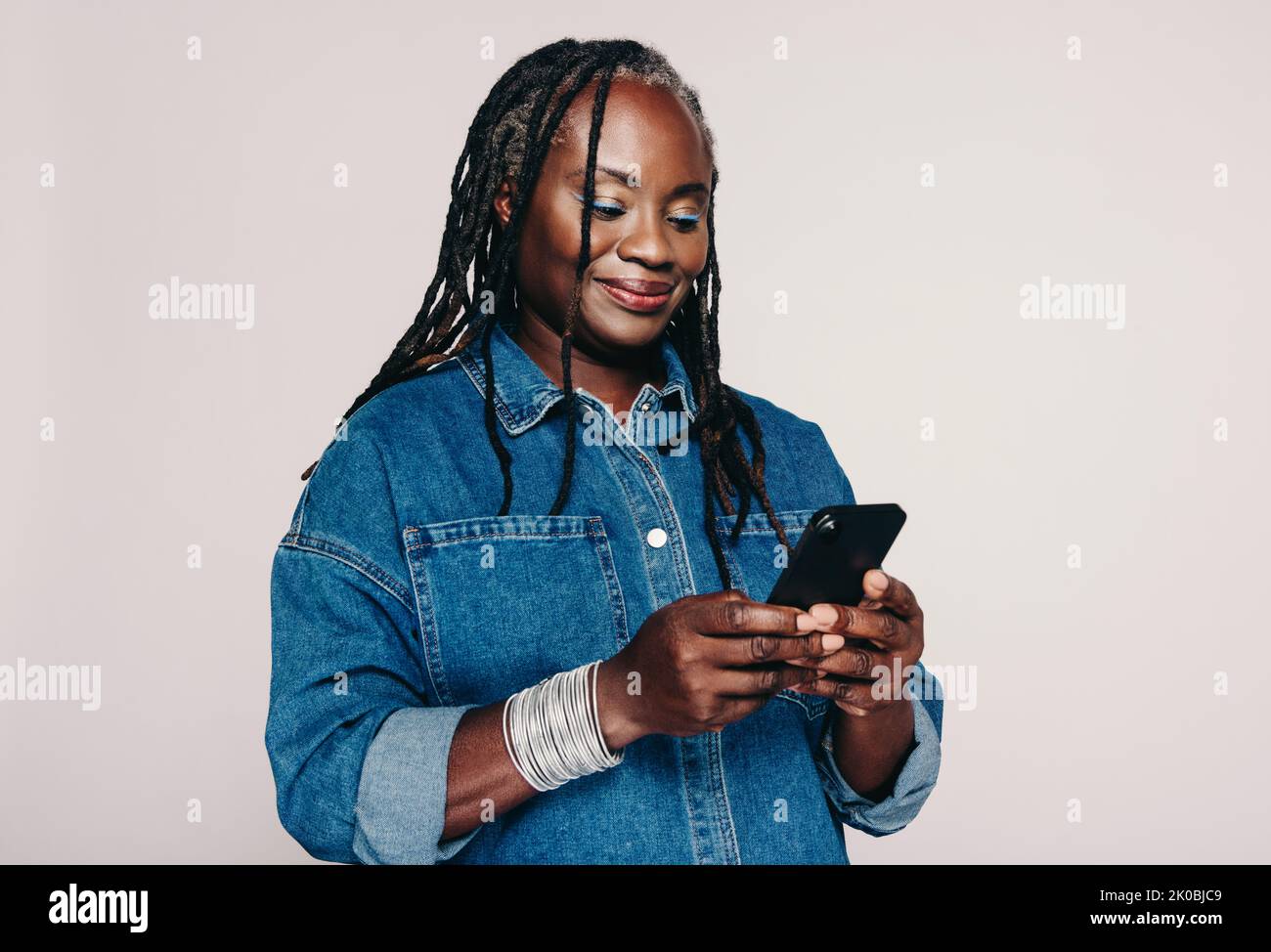 Donna di mezza età che legge un messaggio di testo su uno smartphone mentre si trova su uno sfondo grigio. Donna alla moda con greggi che indossano un denim j Foto Stock